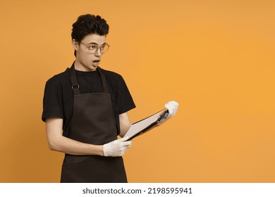 A Young Man In A Black T-shirt And Apron In Work Gloves Holds A Tablet For Papers In His Hands And Looks In Surprise At The Copy Space