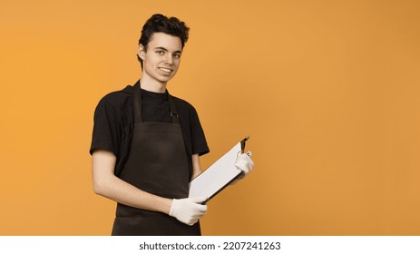 A Young Man In A Black T-shirt And Apron In Work Gloves Holds A Tablet For Papers In His Hands Stands Against A Colored Background With Copy Space