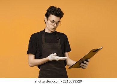 A Young Man In A Black T-shirt And Apron In Work Gloves Is Perplexed And Looks At A Tablet For Papers In His Hands