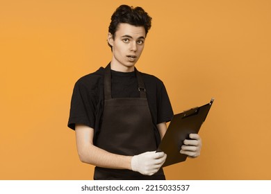 A Young Man In A Black T-shirt And Apron In Work Gloves Looks At The Camera In Surprise And Holds A Tablet For Papers In His Hands