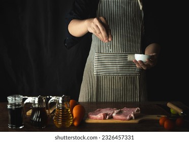 Young man in black uniform and kitchen apron pours sea salt on raw steak. Cooking process.  Horizontal photo with a dark black background. - Powered by Shutterstock