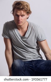 Young Man With Blonde Hair And Blue Eyes, Dressed In Grey T-shirt And Blue Jeans, Sitting On The Stool And Looking At Camera