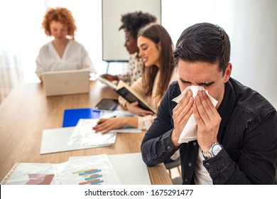 Young Man Blowing His Nose While Working. Young Sick Businessman Blowing His Nose While Working In The Office. There Are People In The Background. Businessman Blowing His Nose With A Tissue At Work