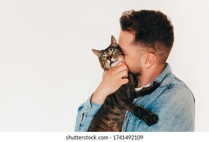 A young man in blue denim shirt is holding and hugging a cute funny brown tabby cat. Home pets. Light background. Pet lover. Copy space for text
