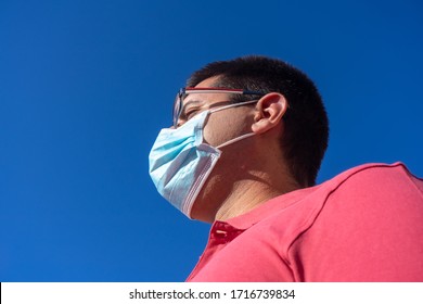 Young Man With Blue Face Mask. He Is Looking At The Sky. He Is Wearing A Salmon Colored Polo Shirt And Glasses.