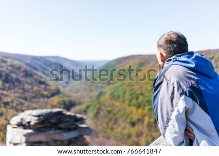 Young man in blue jacket looking at canaan valley mountains in Blackwater falls state park in West Virginia during colorful autumn fall season with yellow foliage on trees, rock cliff at Lindy Point