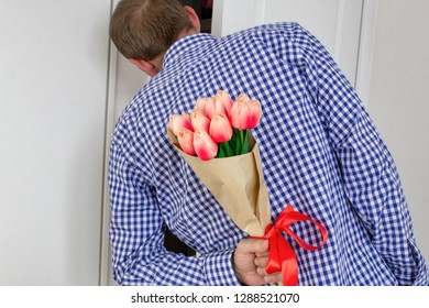 A young man in a blue plaid shirt and jeans, holding a bouquet of tulips behind his back, and peeks in the open door, for white background. Valentine's Day and March 8 concept. - Shutterstock ID 1288521070