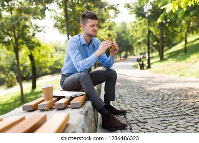 Young Man In Blue Shirt Eating Sandwich With Cup Of Coffee To Go And Laptop Near On Bench In Green City Park