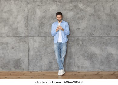 A young man in a blue shirt and jeans stands with his back against a concrete wall. He is using his smartphone and smiling as he looks down at the screen. The floor is made of wood. - Powered by Shutterstock