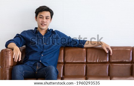 A young man in blue shirt and blue jeans is sitting on a dark brown sofa with smile. He stretch his left arm out for relax.