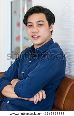 A young man in blue shirt and blue jeans is sitting on a dark brown sofa with smile. He folds his arms in relax gesture.