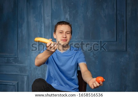 a young man in a blue t-shirt with a banana and a tangerine in his hands, taken by the light of a softbox