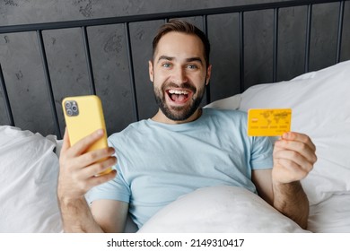 Young Man In Blue T-shirt Lying In Bed Using Mobile Cell Phone Hold Credit Bank Card Shopping Online Book Tour Rest Relax Spend Time In Bedroom Home In Own Room House. Good Mood Night Bedtime Concept