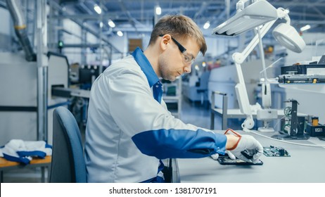 Young Man In Blue And White Work Coat Is Using Plier To Assemble Printed Circuit Board For Smartphone. Electronics Factory Workers In A High Tech Factory Facility.