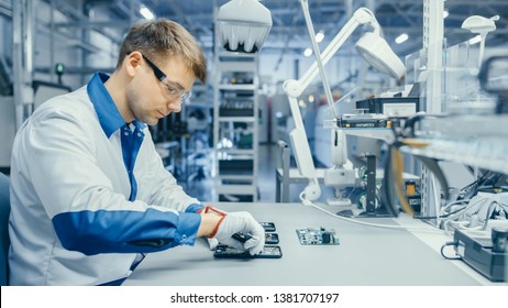 Young Man In Blue And White Work Coat Is Using Plier To Assemble Printed Circuit Board For Smartphone. Electronics Factory Workers In A High Tech Factory Facility.