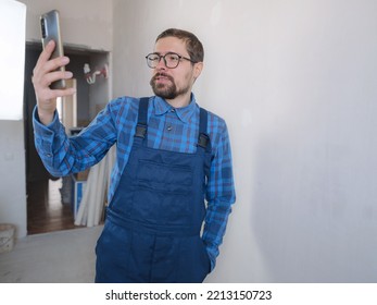 Young Man In Blue Work Suit Doing Repair Apartment. Home Renovation Concept. Man Having Video Call On Smartphone Over White Concrete Wall Background