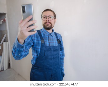 Young Man In Blue Work Suit Doing Repair Apartment. Home Renovation Concept. Man Having Video Call On Smartphone Over White Concrete Wall Background