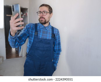 Young Man In Blue Work Suit Doing Repair Apartment. Home Renovation Concept. Man Having Video Call On Smartphone Over White Concrete Wall Background