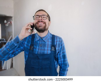 Young Man In Blue Work Suit Doing Repair Apartment. Home Renovation Concept. Man Having Video Call On Smartphone Over White Concrete Wall Background