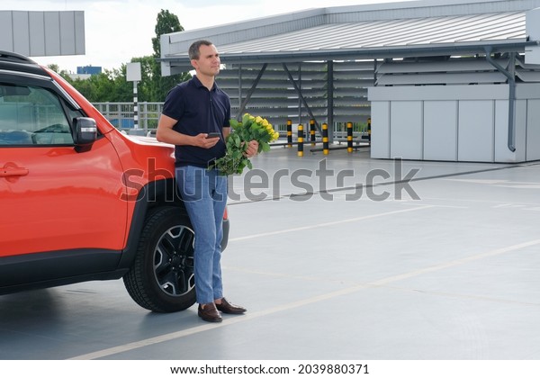 Young man with a bouquet of flowers stands near\
the car in the parking