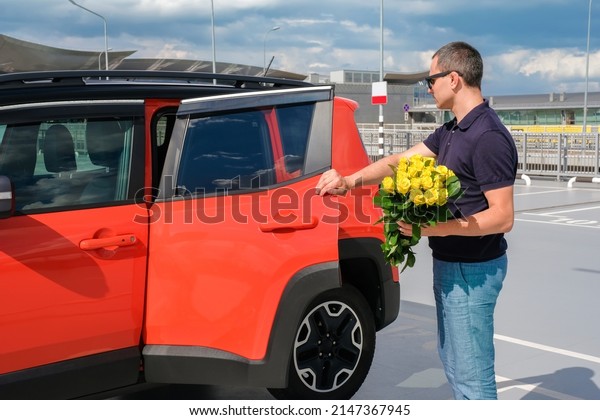 A young man with a\
bouquet of flowers stands near his car in the parking lot.\
Vacation, business trip