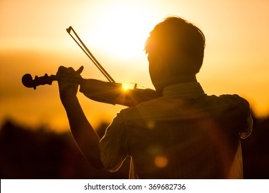 Young man or boy playing the violin at sunset standing with his back to the camera with his instrument and bow silhouetted against the fiery orange sky