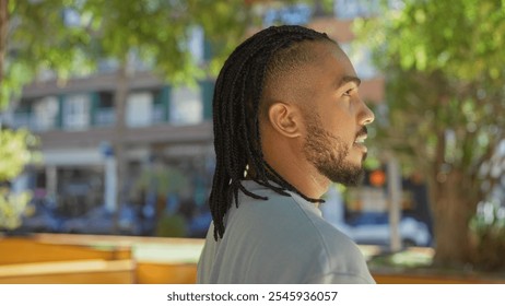 Young man with braids standing in a sunny urban park, showcasing a relaxed and thoughtful expression outdoors amidst vibrant greenery and modern city buildings. - Powered by Shutterstock