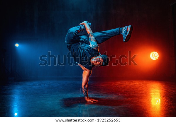 Young man break dancing in dark hall with blue and\
red lights. Tattoo on\
hands.