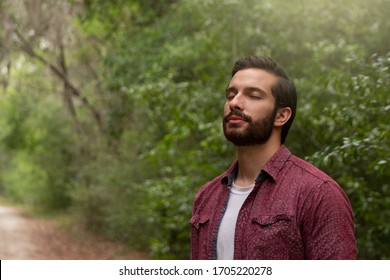 young man with brown hair and beard wearing red button down breathes in a breath of fresh air immersing himself in a nature path to social distance himself in order to stay healthy and alive.