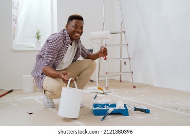 A Young Man In Brown Jeans And A Red Plaid Shirt Picks Up A Can Of White Paint And Pours It On A Tray. He Is Renovating One Of The Rooms In The House, Painting The Walls.