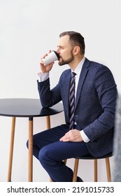 A Young Man In A Business Suit At A Small Round Table Drinking Coffee Or Tea From A White Cup. A Young Businessman Is Relaxing Over A Cup Of Coffee.