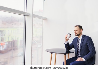 A Young Man In A Business Suit At A Small Round Table Drinking Coffee Or Tea From A White Cup. A Young Businessman Is Relaxing Over A Cup Of Coffee.