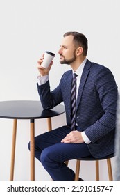 A Young Man In A Business Suit At A Small Round Table Drinking Coffee Or Tea From A White Cup. A Young Businessman Is Relaxing Over A Cup Of Coffee.