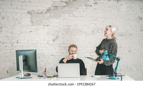 Young Man And Business Woman Laughing At Contemporary Office Space Besides White Desk With Computer Laptop Lamp Pencils Notebooks And White Brick Wall Background