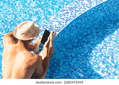 Young man businessman working online during summer vacation using digital device mobile phone in resort hotel poolside. Guy hold smartphone isolated on blue water swimming pool background copy space - Powered by Shutterstock