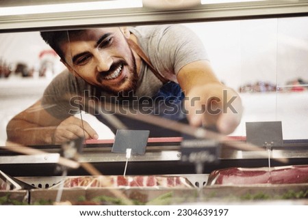 Young man butcher arranging meat products in display case of butcher shop