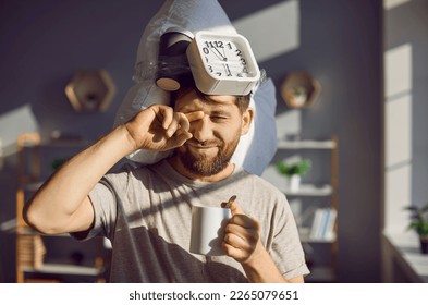 Young man can't wake up for work very early in the morning. Portrait of a man with a pillow and an alarm clock on his head holding his coffee cup and rubbing his sleepy eyes in the bright sun