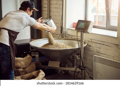 Young man in cap pouring green coffee beans into metal hopper while using electronic scales
