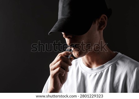 A young man with a cap is vaping an e-cigarette, highlighting the risks of nicotine addiction in a moody atmosphere.