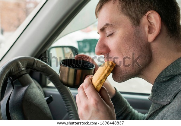 A young man in a car behind\
the wheel eats a pie with pleasure and holds a metal mug in his\
hand