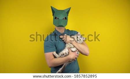 A young man in a cardboard jackal mask holds a cat in his arms on a yellow background. Man in a green polo and mask.