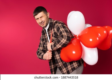 A Young Man Carries A Bunch Of Heart-shaped Balloons And Squints At The Camera. St.Valentine's Day On Red Background. High Quality Photo