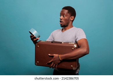 Young Man Carrying Suitcase And Running Late For Vacation Trip, Looking At Time On Clock Over Blue Background. Person With Retro Briefcase Having Watch To Prepare For Work Voyage.