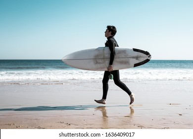 Young man carrying surfboard and running on sunny beach. Handsome guy wearing wetsuit with ocean in background. Surfboarding concept. Side view. - Powered by Shutterstock