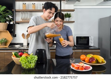 Young man in casual clothe pour mixed fruit and veggie smoothie from blending machine into his girlfriend glass. Asian lover spend  morning time together in the kitchen. 