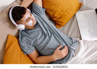 Young Man With Casual Clothes And Beauty Mask On The Face Lying On Bed With Laptop Computer And Resting. Cosmetic For Men, Skin Care. Man Listening Music Or Audio Book. Relaxing At Home.