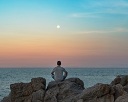 Young Man In Casual Clothes Sits On Rocks By The Sea At Sunset, Doing Meditation  And Looking At The Moon Over His Head