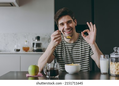 Young Man In Casual Clothes Striped T-shirt Eat Breakfast Muesli Cereals With Milk Fruit In Bowl Sit By Table Show Ok Gesture Cooking Food In Light Kitchen At Home Alone Health Diet Lifestyle Concept.