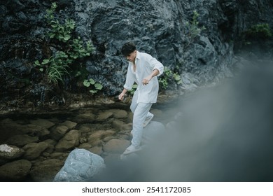 A young man in casual clothing carefully stepping on rocks in a peaceful stream, surrounded by lush greenery and rocky terrain. - Powered by Shutterstock