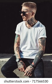 Young man in casual outfit and sunglasses posing at street showing tattoos all over body.
